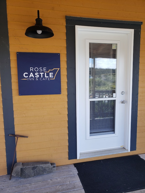 A hotel entrance with a sign reading 'ROSE CASTLE INN & CAFE,' a door with a window, and a black wall-mounted light fixture. - Rose Castle Inn & Café. Beautiful, breathtaking views of the ocean. (Bell Island)