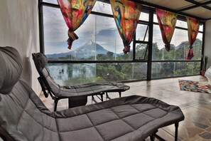 A room with a large window, a sofa, and a view of a mountain. - Aura Arenal: Fire Cabin - Lake & Volcano View (El Castillo)