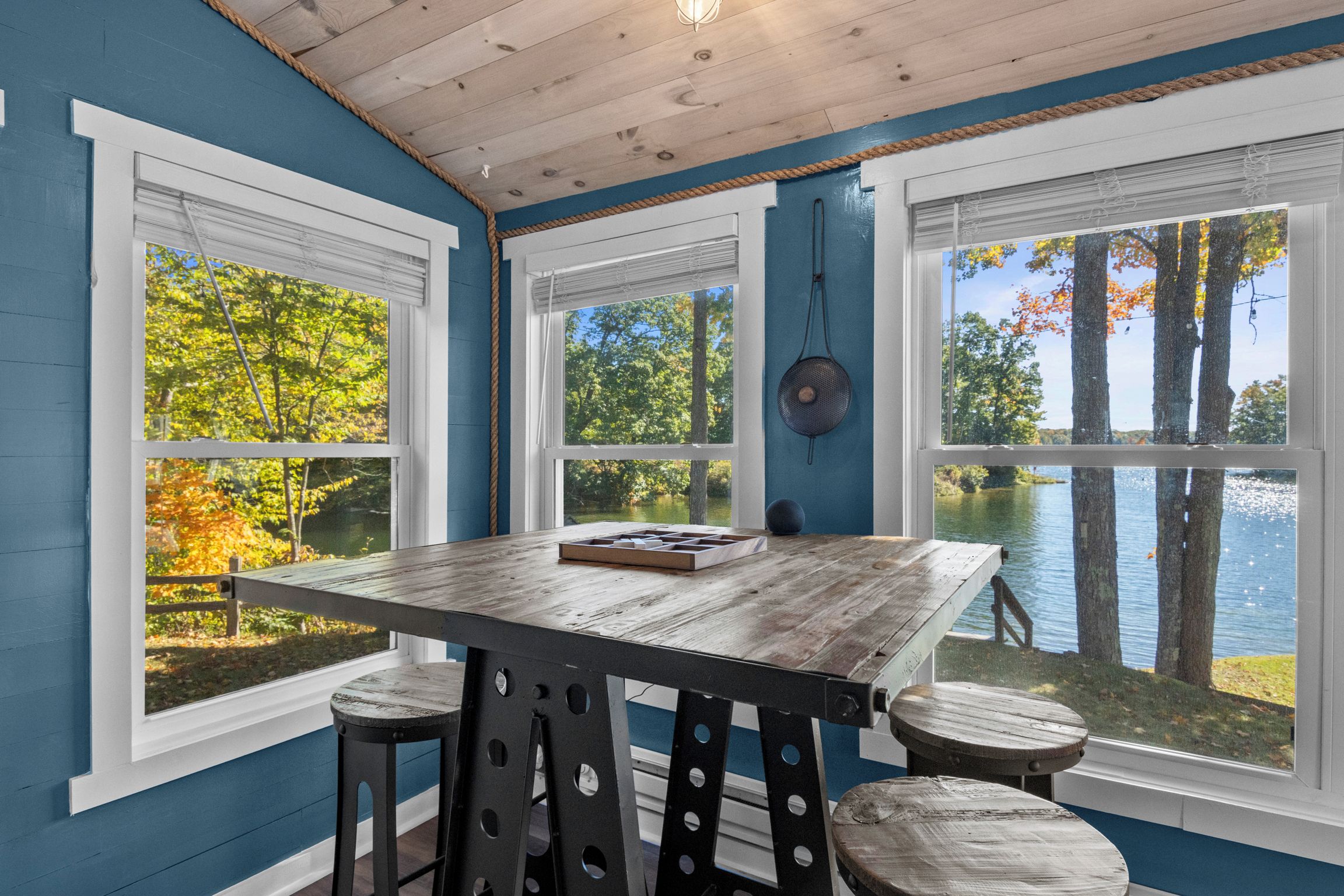 A room with a wooden table and stools, large windows with views of trees and a lake.