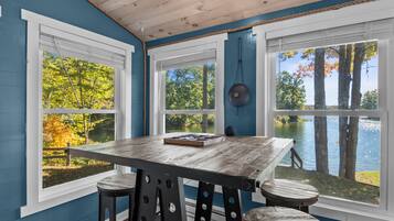 A room with a wooden table and stools, large windows with views of trees and a lake.