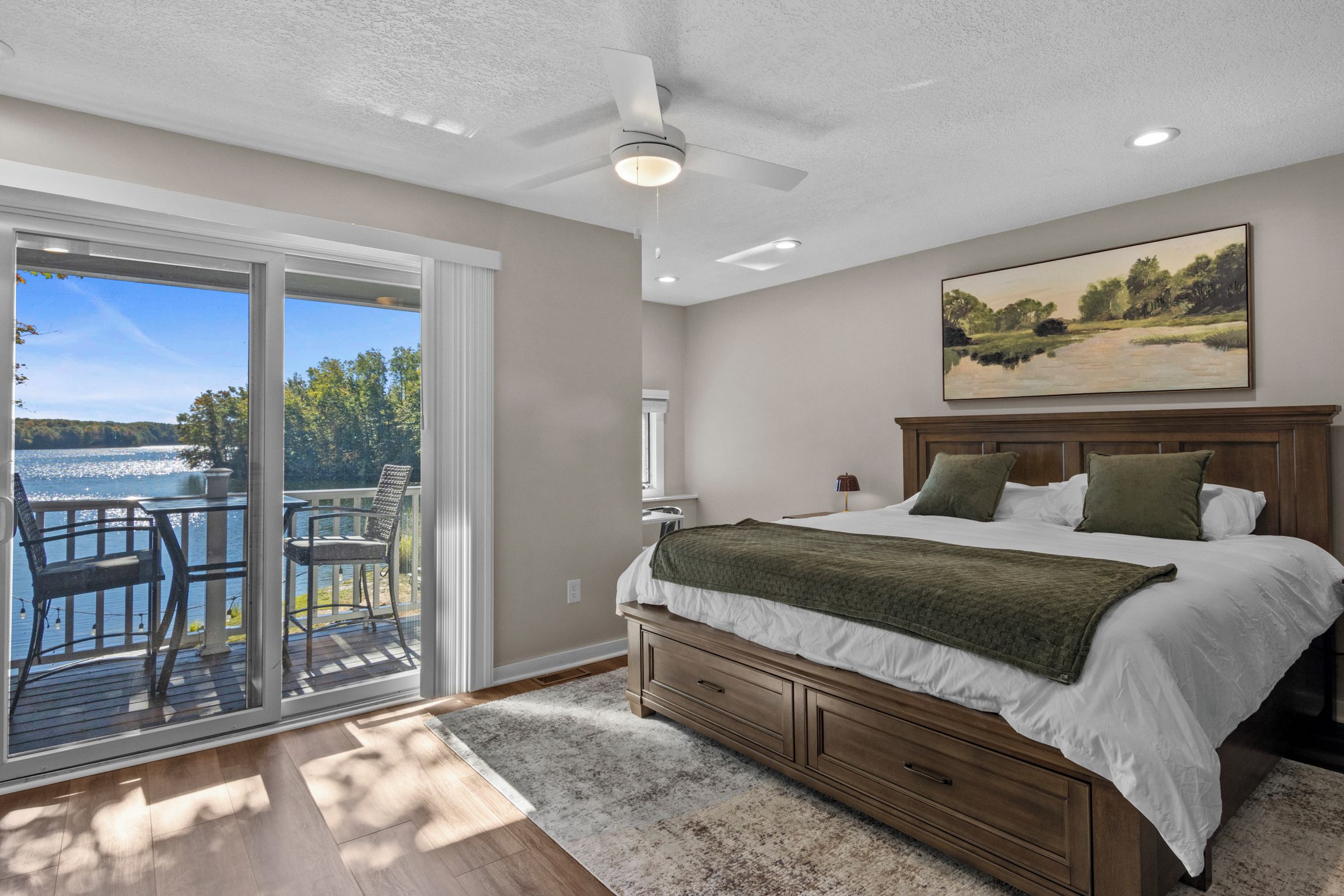 A bedroom with a large bed, a nightstand, a painting, and a view of a lake through a sliding glass door.