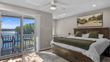 A bedroom with a large bed, a nightstand, a painting, and a view of a lake through a sliding glass door.