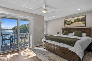Un dormitorio con una cama grande, una mesita de noche, un cuadro y vistas a un lago a través de una puerta corrediza de vidrio.