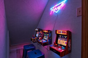 A room with three arcade machines, two blue stools, and a neon sign on the wall.