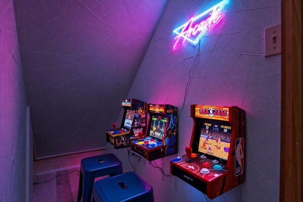 Una habitación con tres máquinas de videojuegos, dos taburetes azules y un letrero de neón en la pared.