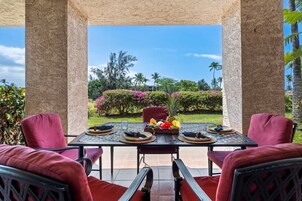 Un área de comedor al aire libre con una mesa dispuesta para cuatro, rodeada de sillas, bajo una estructura cubierta con vista a un jardín y palmeras.