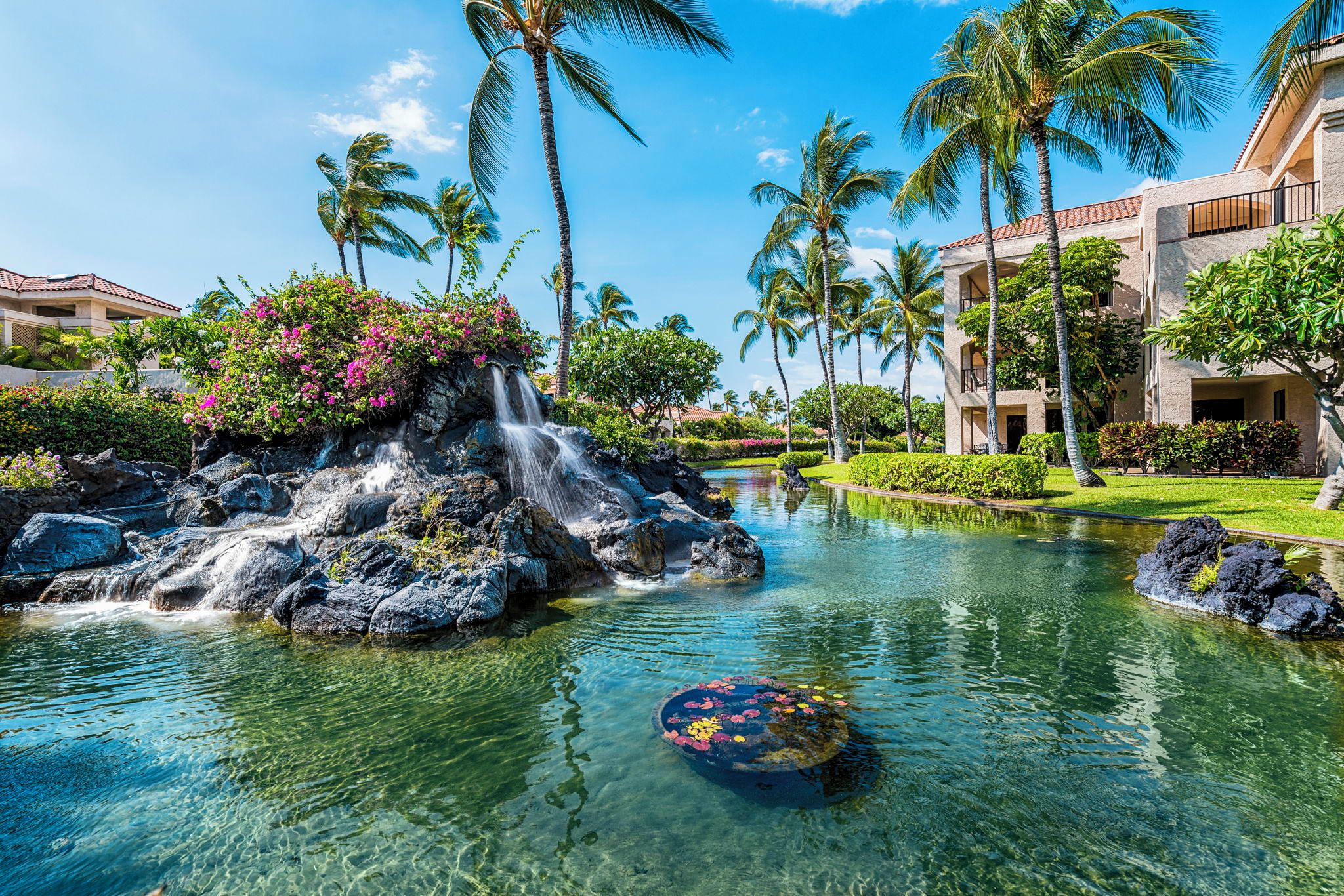 A resort with a clear pool, palm trees, and a waterfall.