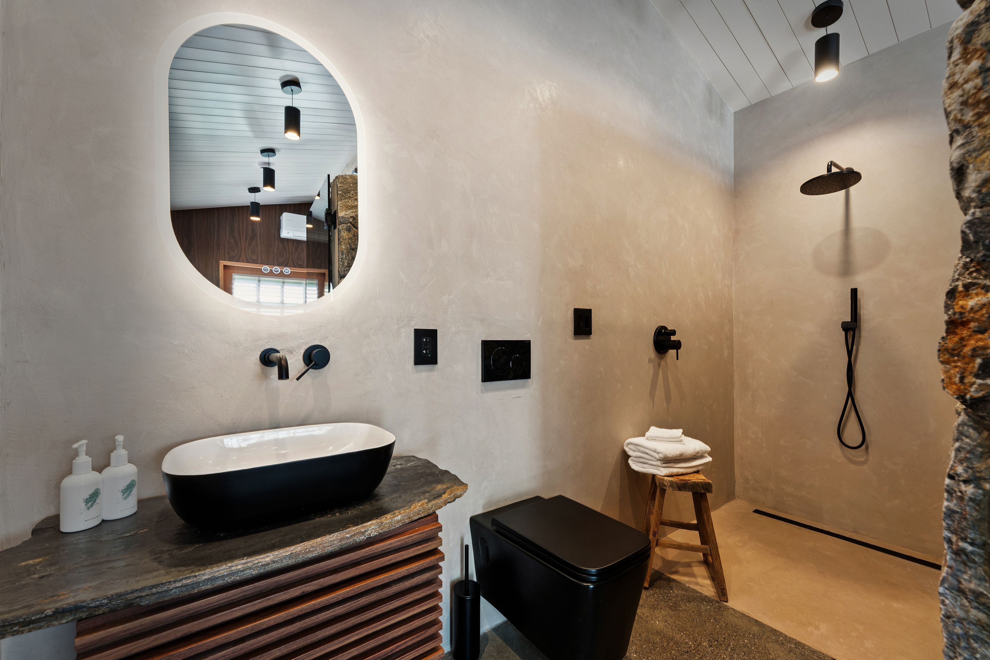 Un baño con un espejo ovalado grande, un lavamanos negro, una ducha y un taburete pequeño.
