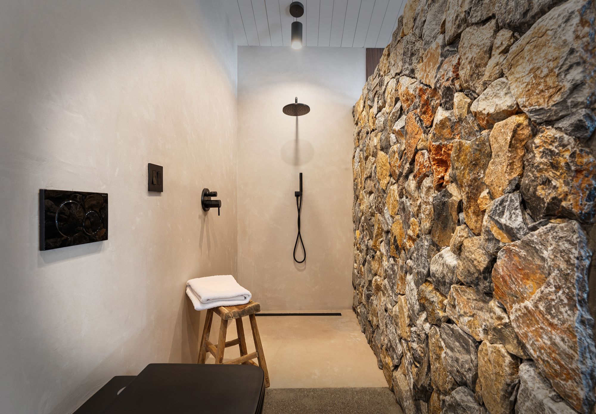 Un baño con pared de piedra, un taburete de madera con una toalla y una ducha con cabezal de ducha manual.