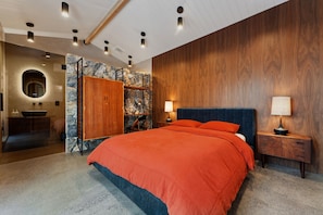 A modern bedroom with a large bed, a wooden headboard, a nightstand, and a stone wall.