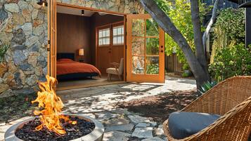 Pared exterior de piedra, un dormitorio con cama y mesita de noche, una silla de mimbre y un hoyo de fuego.