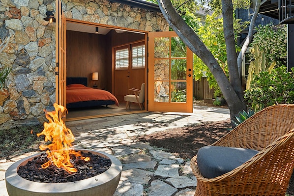 A stone exterior wall, a bedroom with a bed and a nightstand, a wicker chair, and a fire pit.