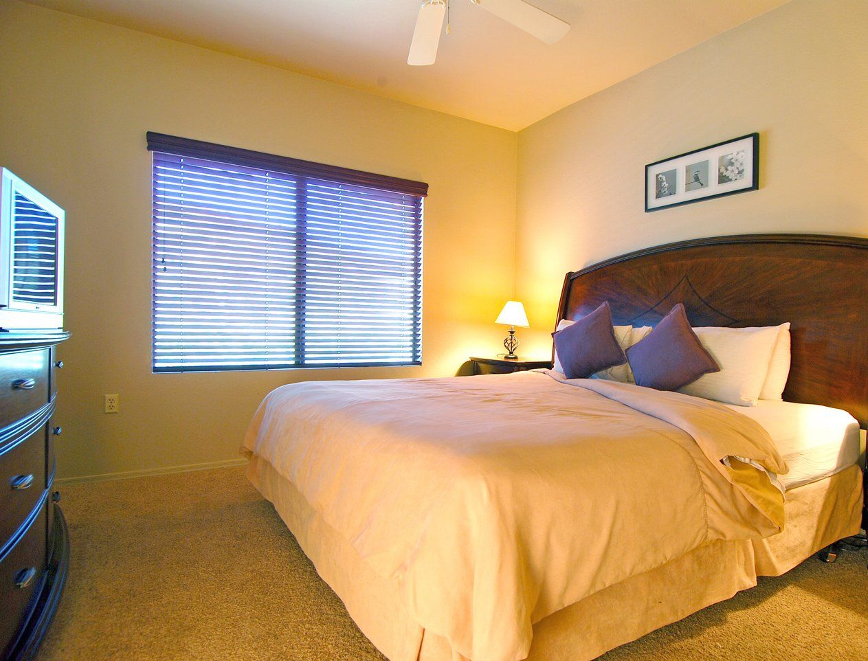 A bedroom with a bed, a dresser, a nightstand with a lamp, and a window with blinds.