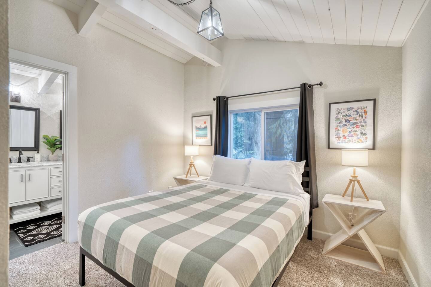 Un dormitorio con cama, mesitas de noche, ventana con cortinas, un cuadro enmarcado y un baño con espejo y lavabo.