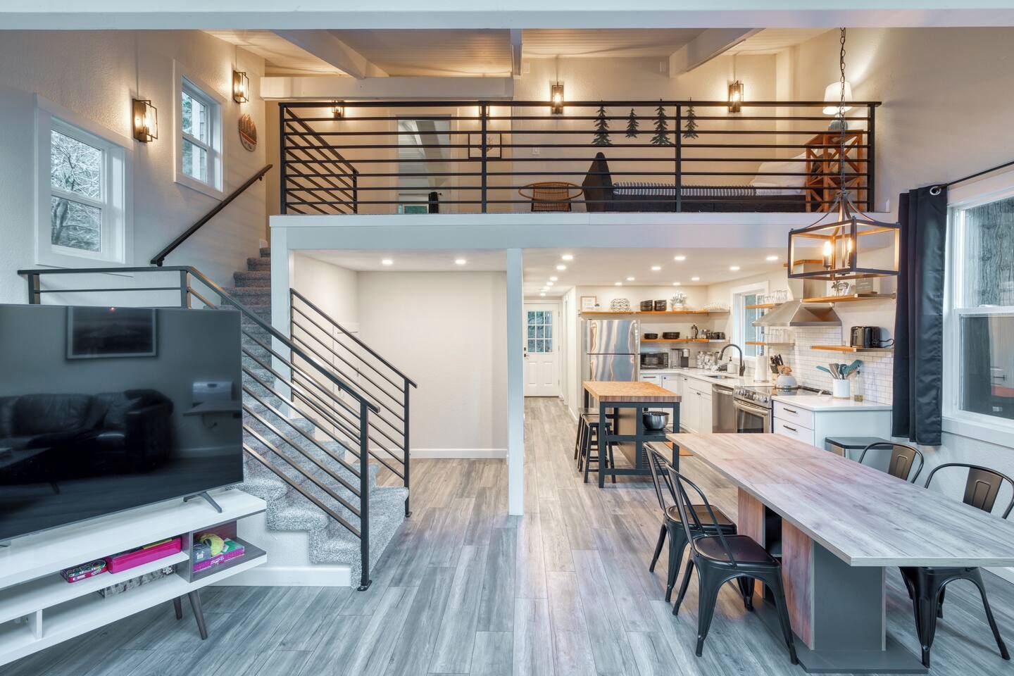 A modern interior with a staircase leading to a loft bedroom, a dining area with a wooden table, and a kitchen with white cabinets.