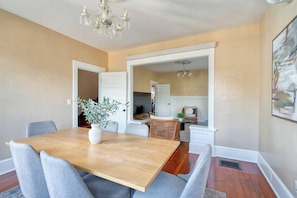 A dining area with a wooden table, grey upholstered chairs, a white vase with greenery, and a chandelier. - Your Front Door Opens onto Mississippi Ave (Portland)