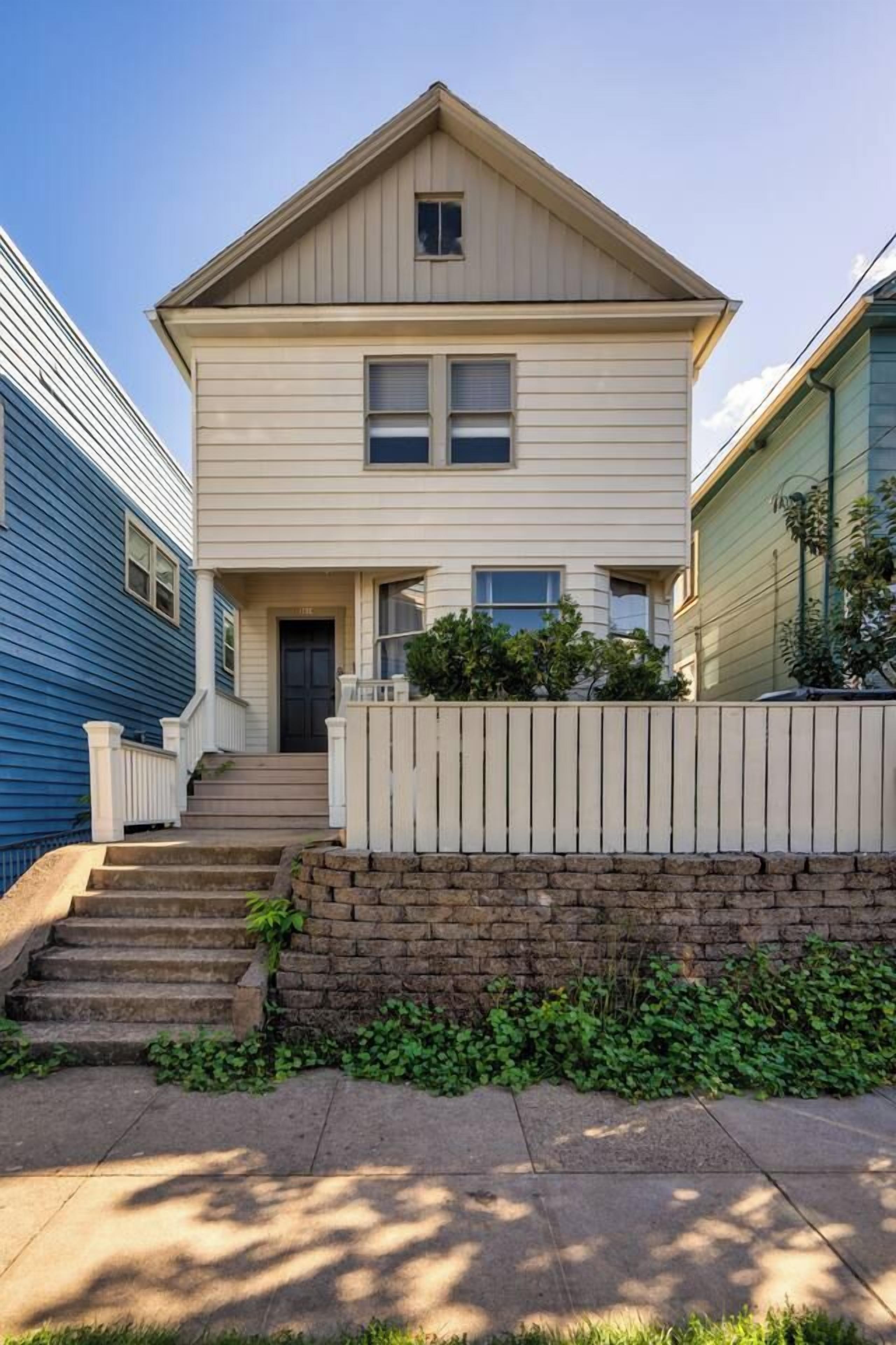 A two-story residential building with a front porch, a brick foundation, and a small front yard.