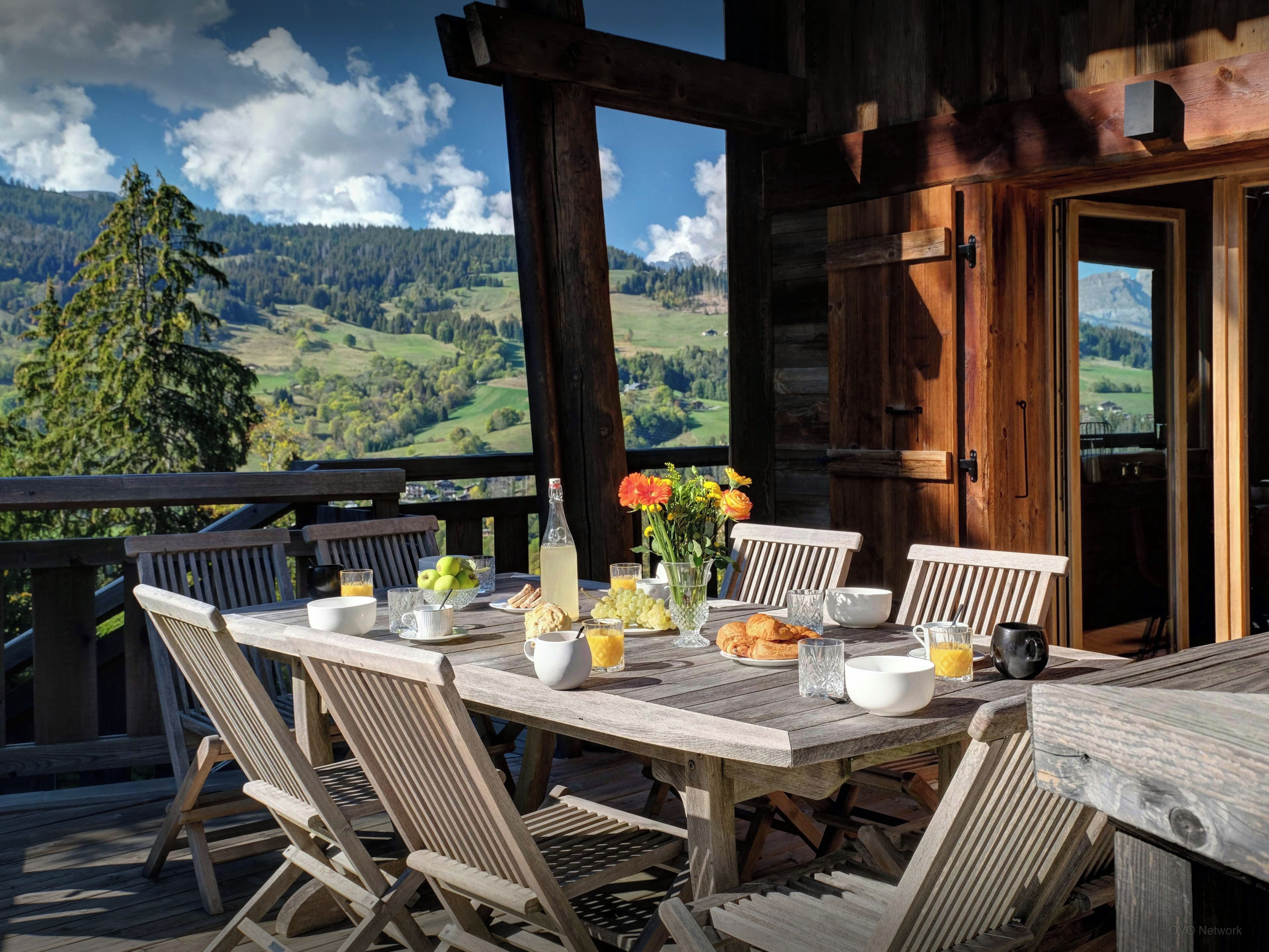 Une terrasse en bois avec une table dressée pour le petit-déjeuner, offrant une vue sur un paysage montagneux.