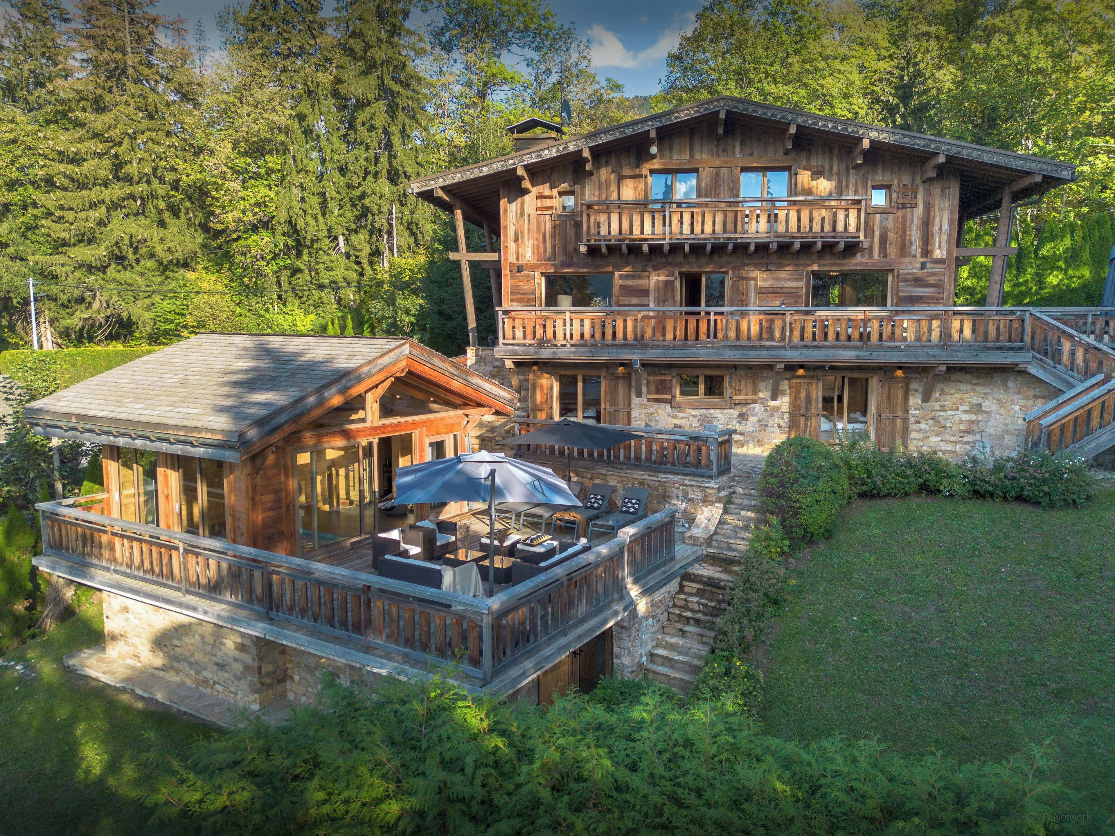 Une cabane en bois à deux étages, avec des fondations en pierre, un balcon et un patio aménagé avec du mobilier d’extérieur.