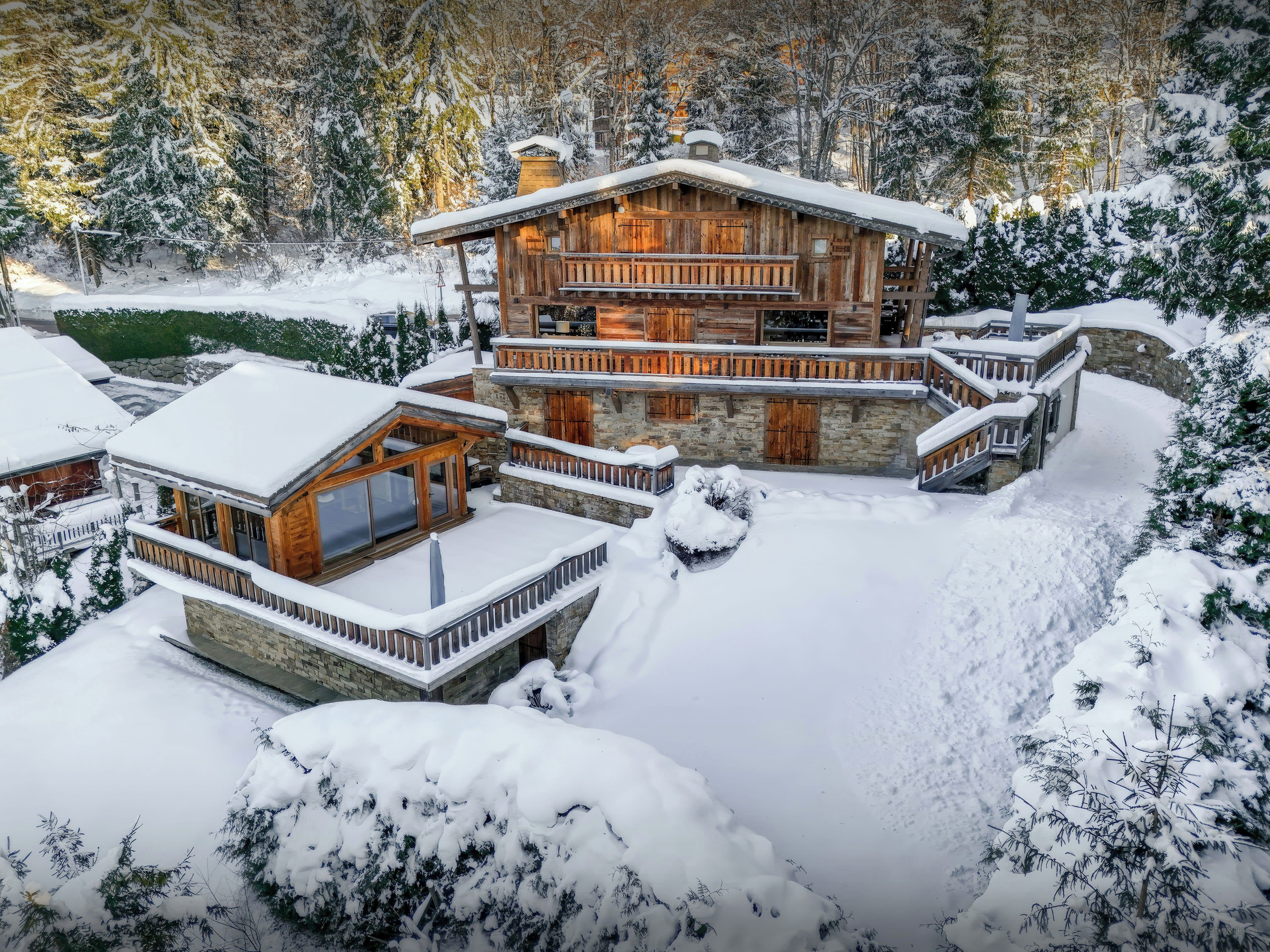 Une cabane en bois à plusieurs niveaux, avec des fondations en pierre, entourée d’arbres couverts de neige.