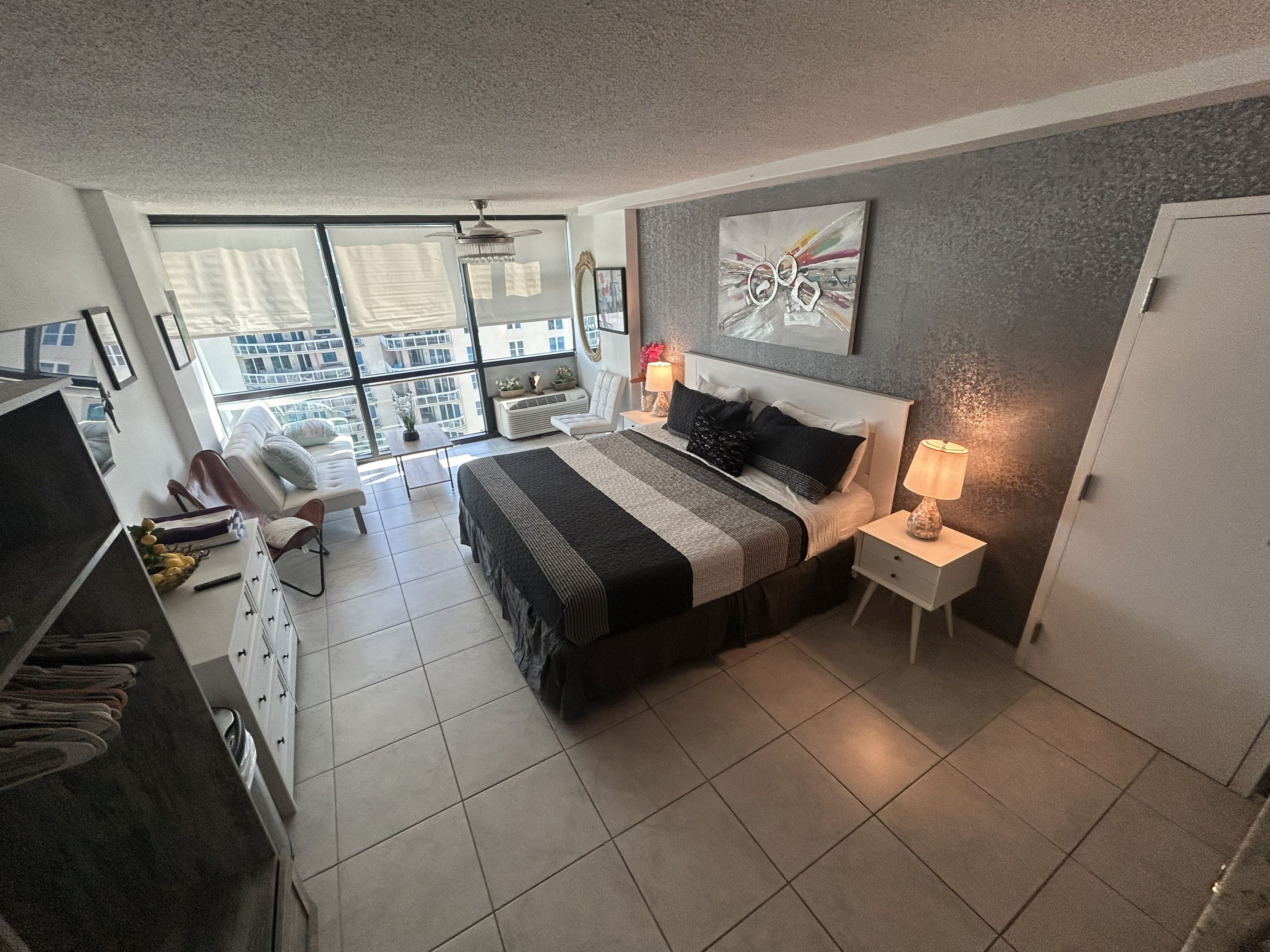 Une chambre moderne avec un grand lit, un bureau et une chaise, une grande fenêtre donnant sur des immeubles et un ventilateur de plafond.