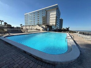 Een zwembad met een helderblauw wateroppervlak, omgeven door een geplaveid terras. Op de achtergrond staat een groot hotelgebouw met meerdere verdiepingen en een glazen gevel.