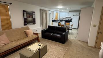 A living room with a sofa, a coffee table, and a rug. There is a kitchen in the background with cabinets and a refrigerator.