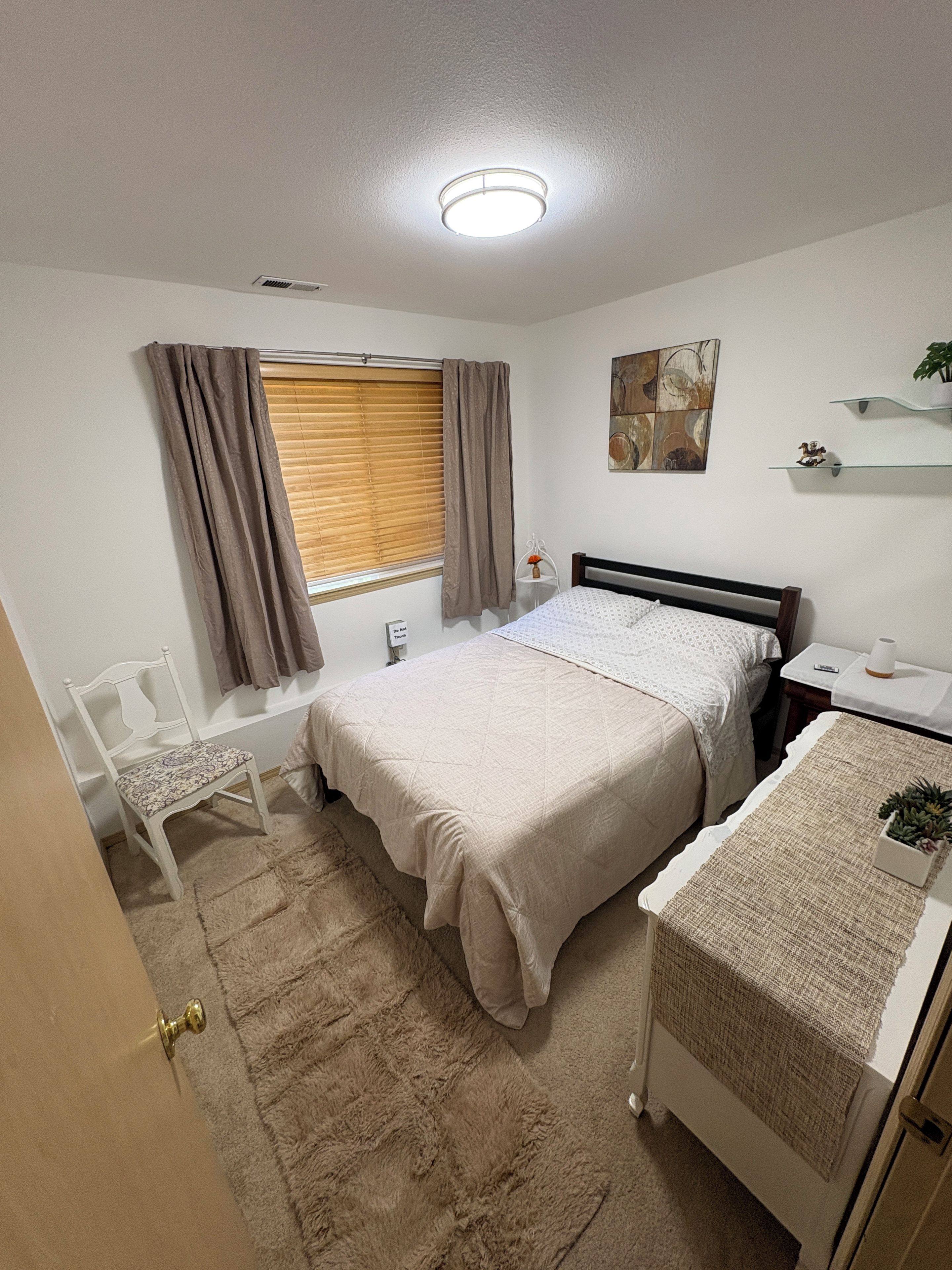 A bedroom with a bed, bedside table, chair, window with curtains, and a ceiling light.