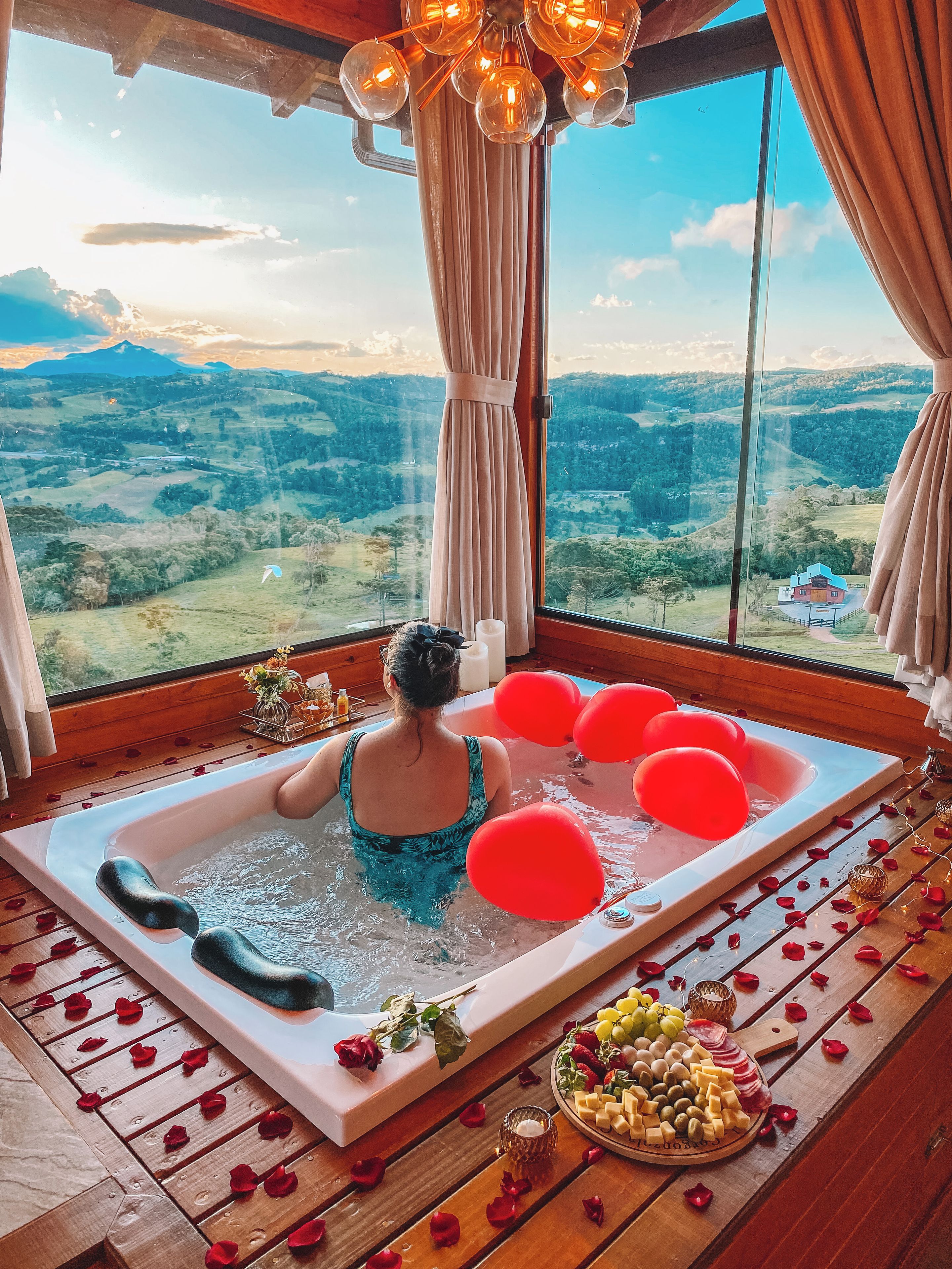 Una habitación con un ventanal grande, una bañera con globos rojos y una mesa con comida y flores.