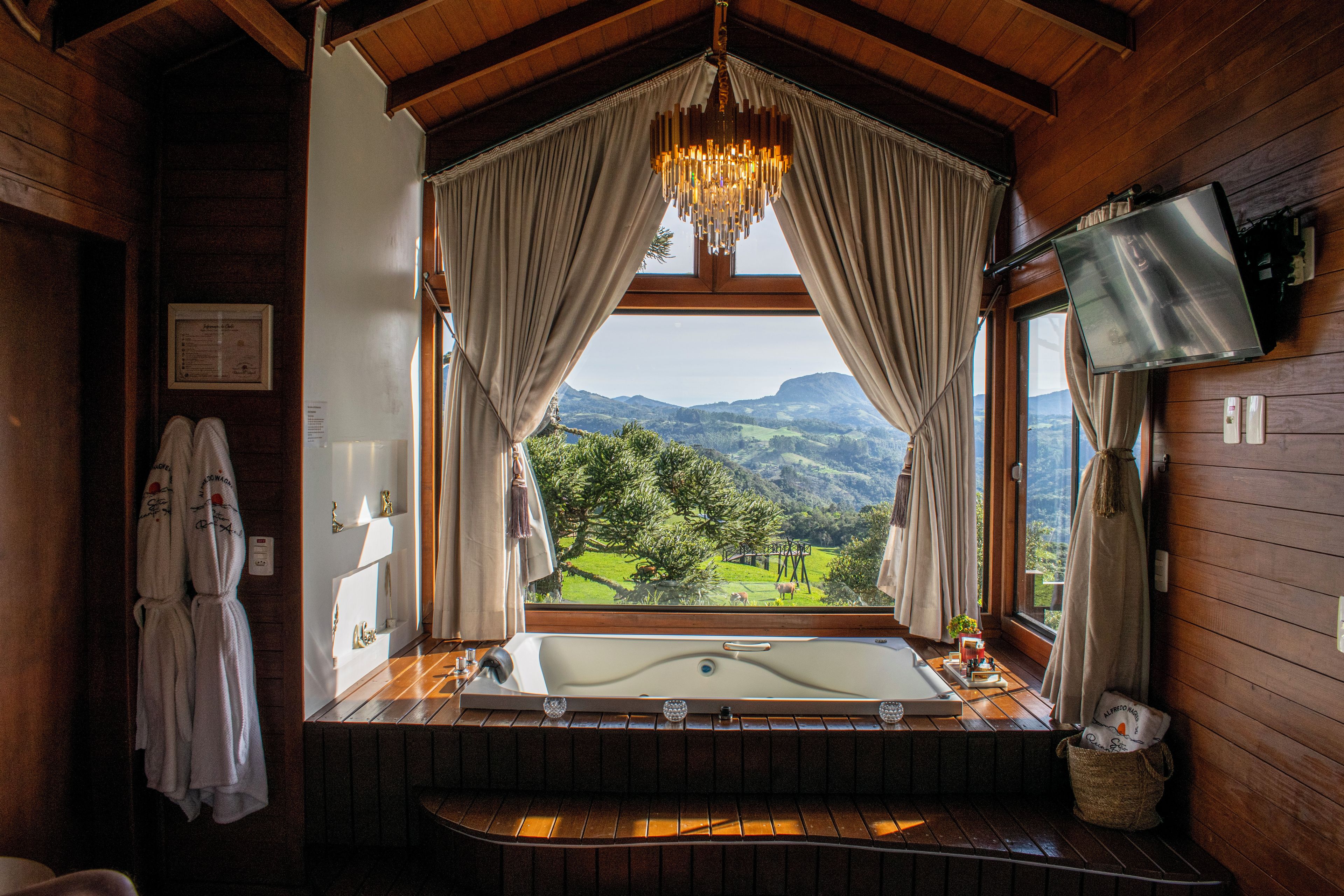 A bathroom with a large bathtub, a chandelier, and a view of a green landscape through a window.