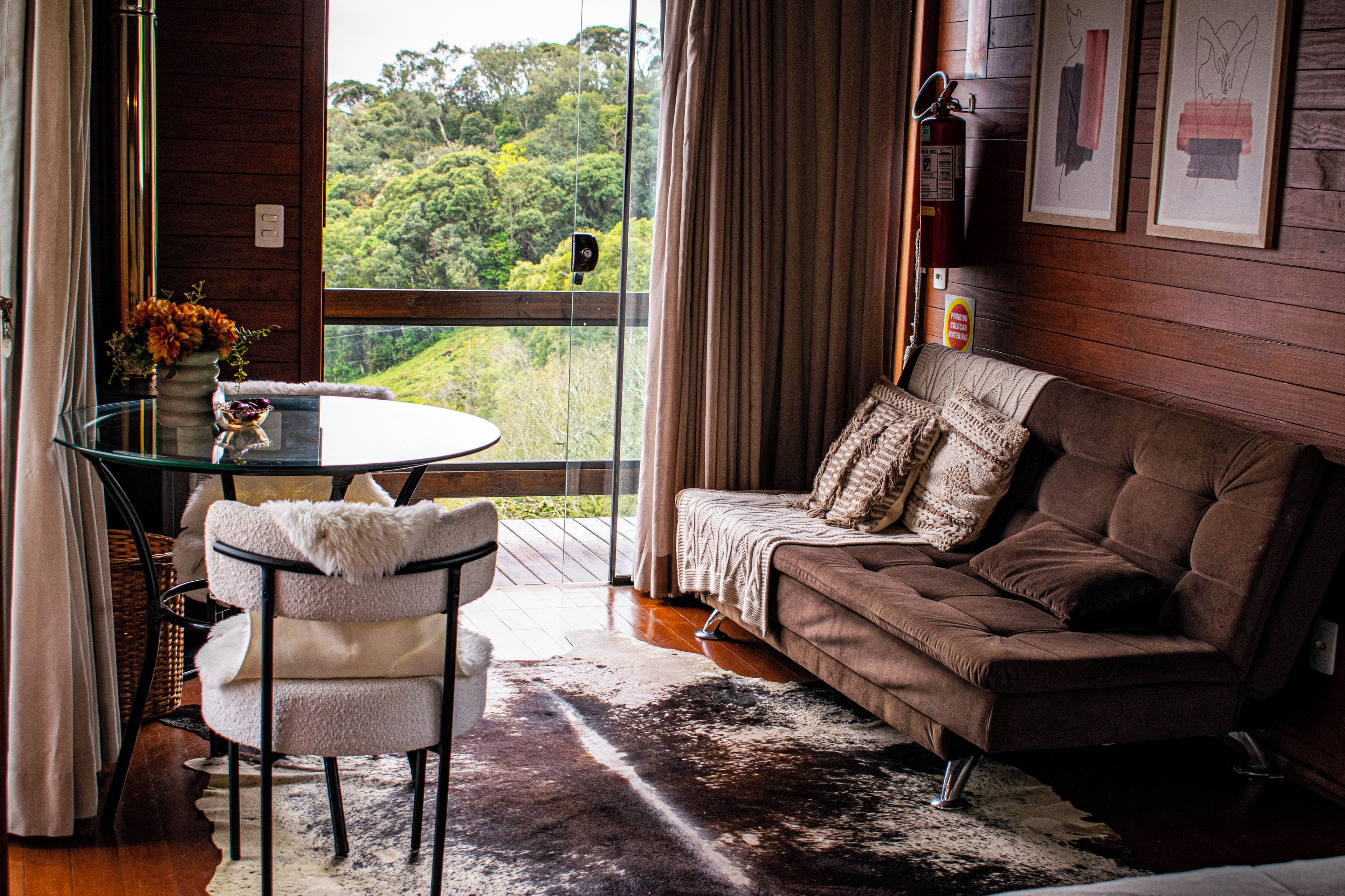 Ein gemütliches Wohnzimmer mit einem Glastisch, einem braunen Sofa und Blick auf einen Wald durch große Fenster.