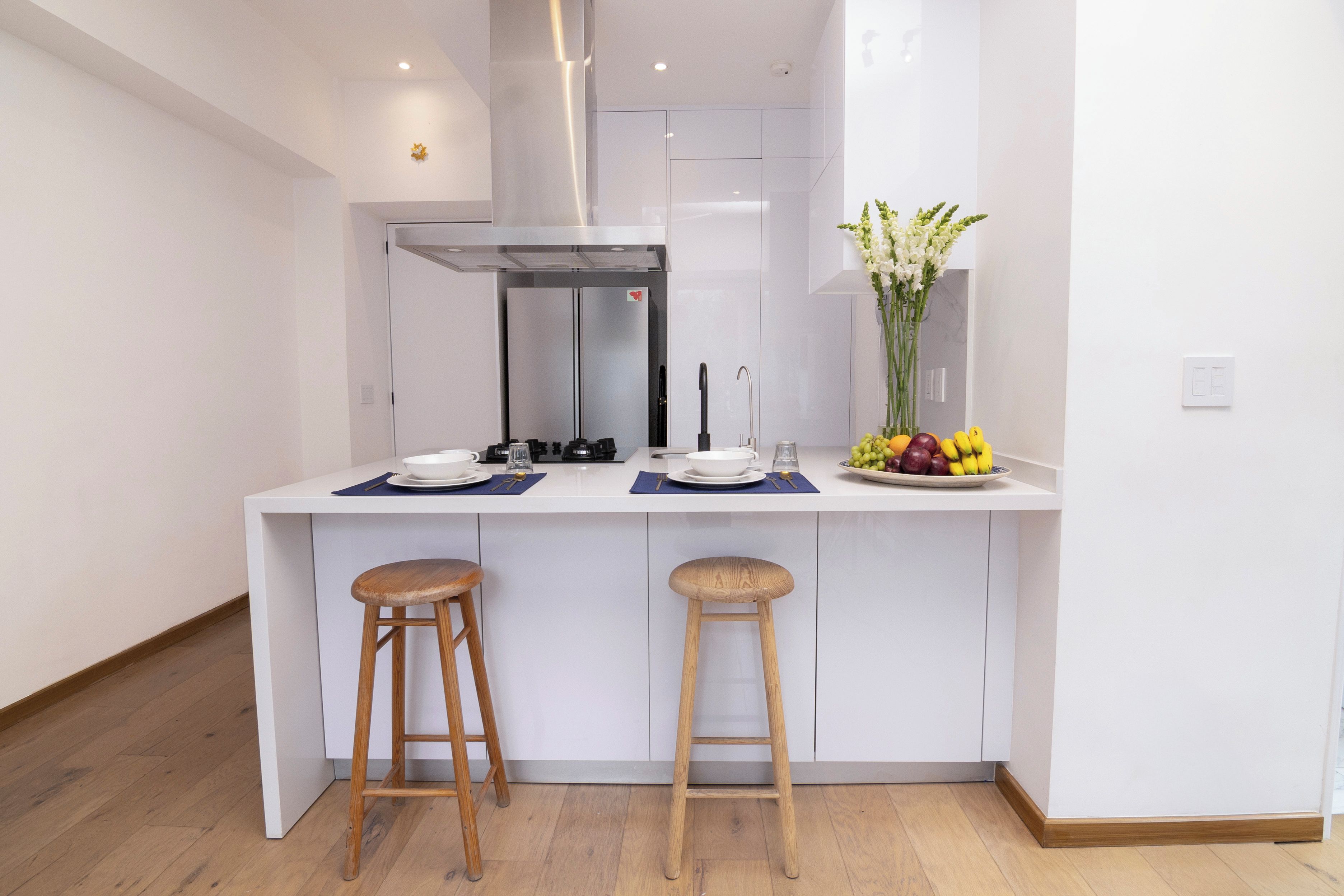 Una cocina moderna con armarios blancos, una campana extractora de acero inoxidable, un lavamanos integrado y dos taburetes de madera.