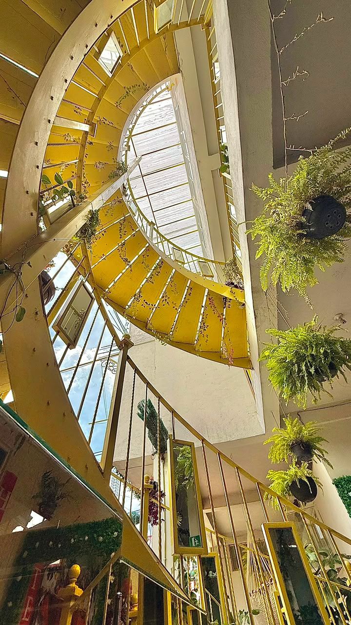 Una scala a chiocciola con ringhiere gialle all'interno di un edificio, circondata da vegetazione e parapetti in vetro.