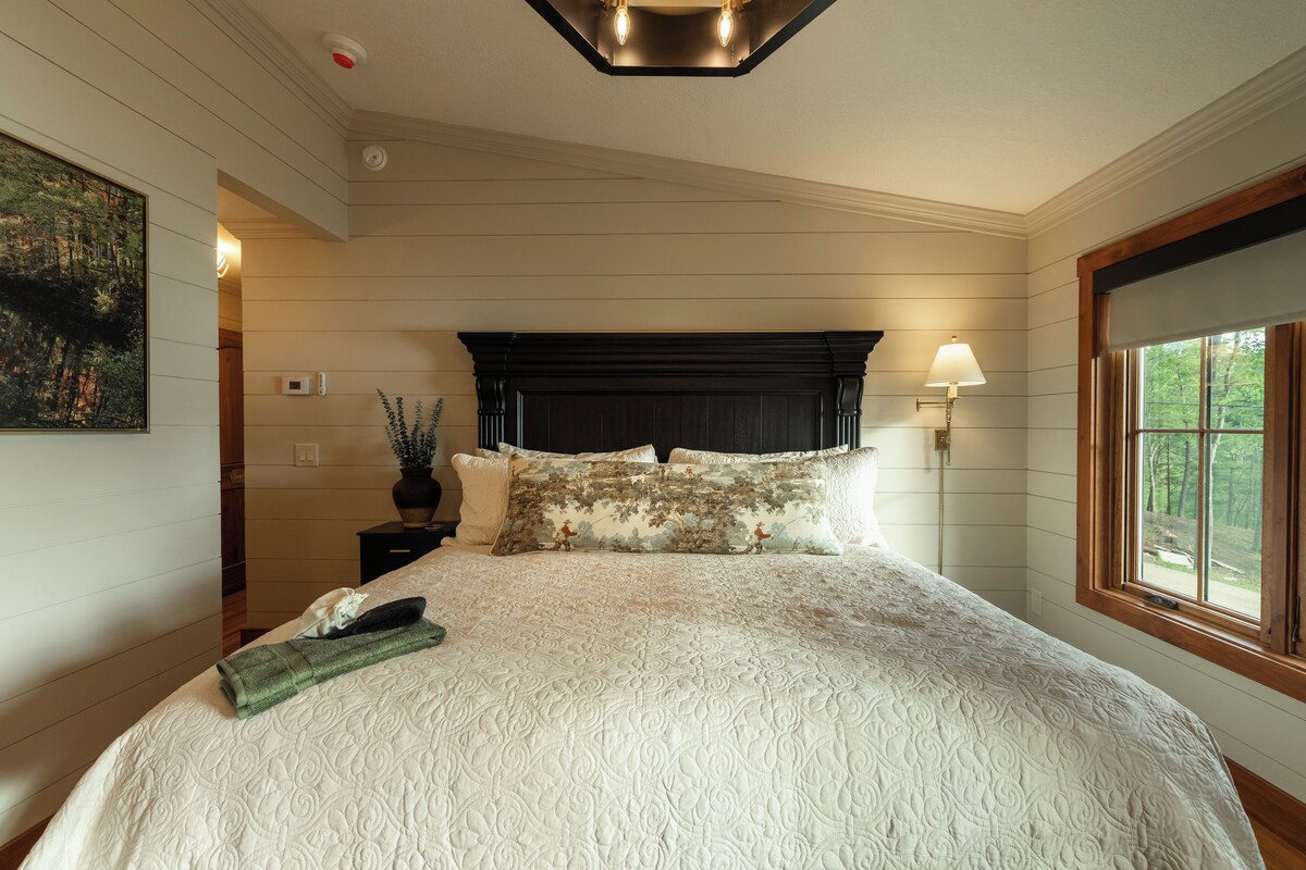 A bedroom with a large bed, a nightstand, a wall-mounted lamp, and a window with a view of trees.