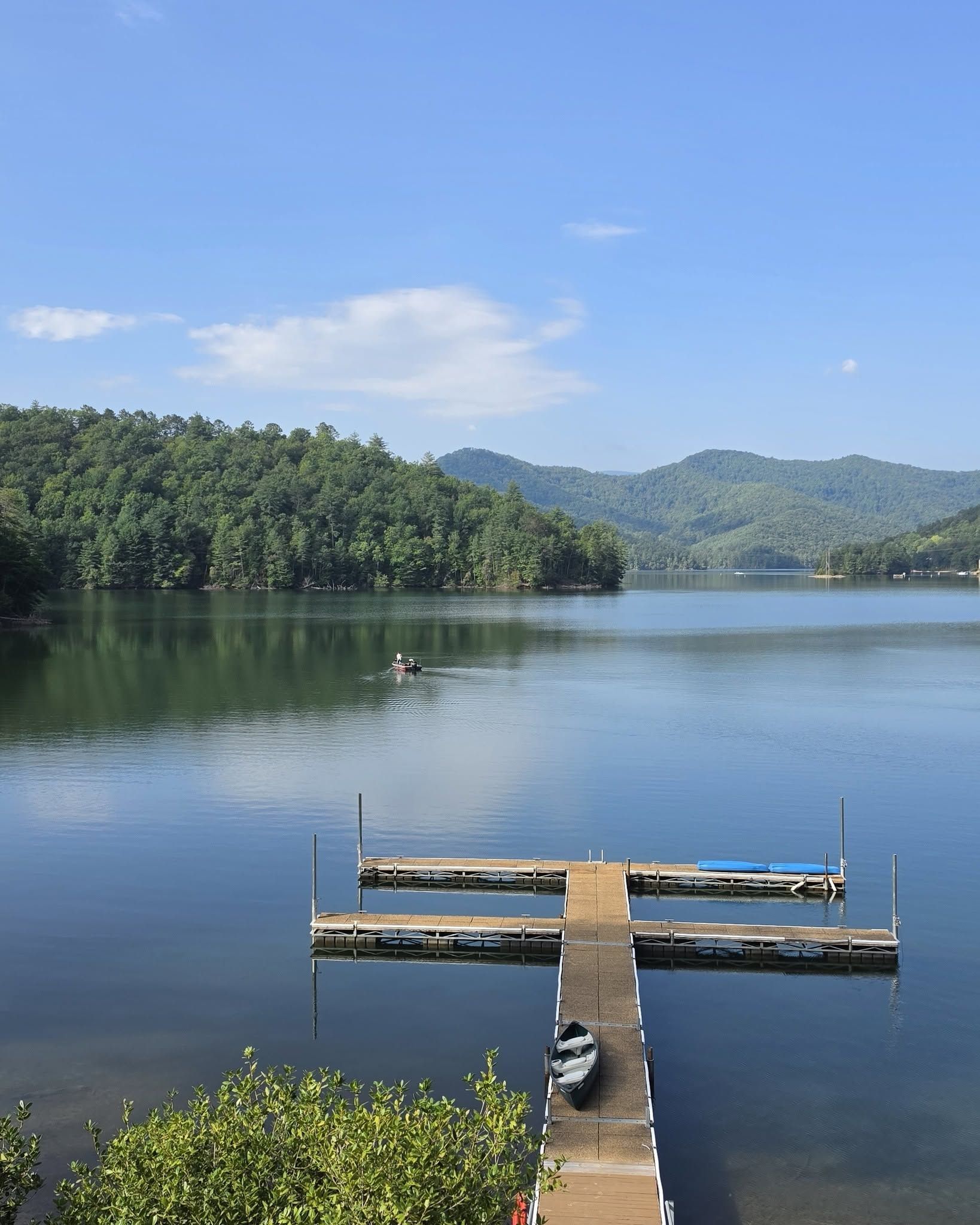 Ein ruhiger See mit einer Holzsteganlage, die ins Wasser ragt, umgeben von dichtem Wald und Hügeln im Hintergrund.