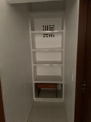 A narrow hallway with a white shelving unit against the wall, a wooden door on the right, and a tiled floor.