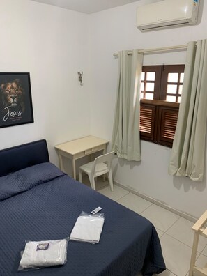 A bedroom with a bed, a desk, a chair, a window with curtains, an air conditioner, and a framed picture on the wall.