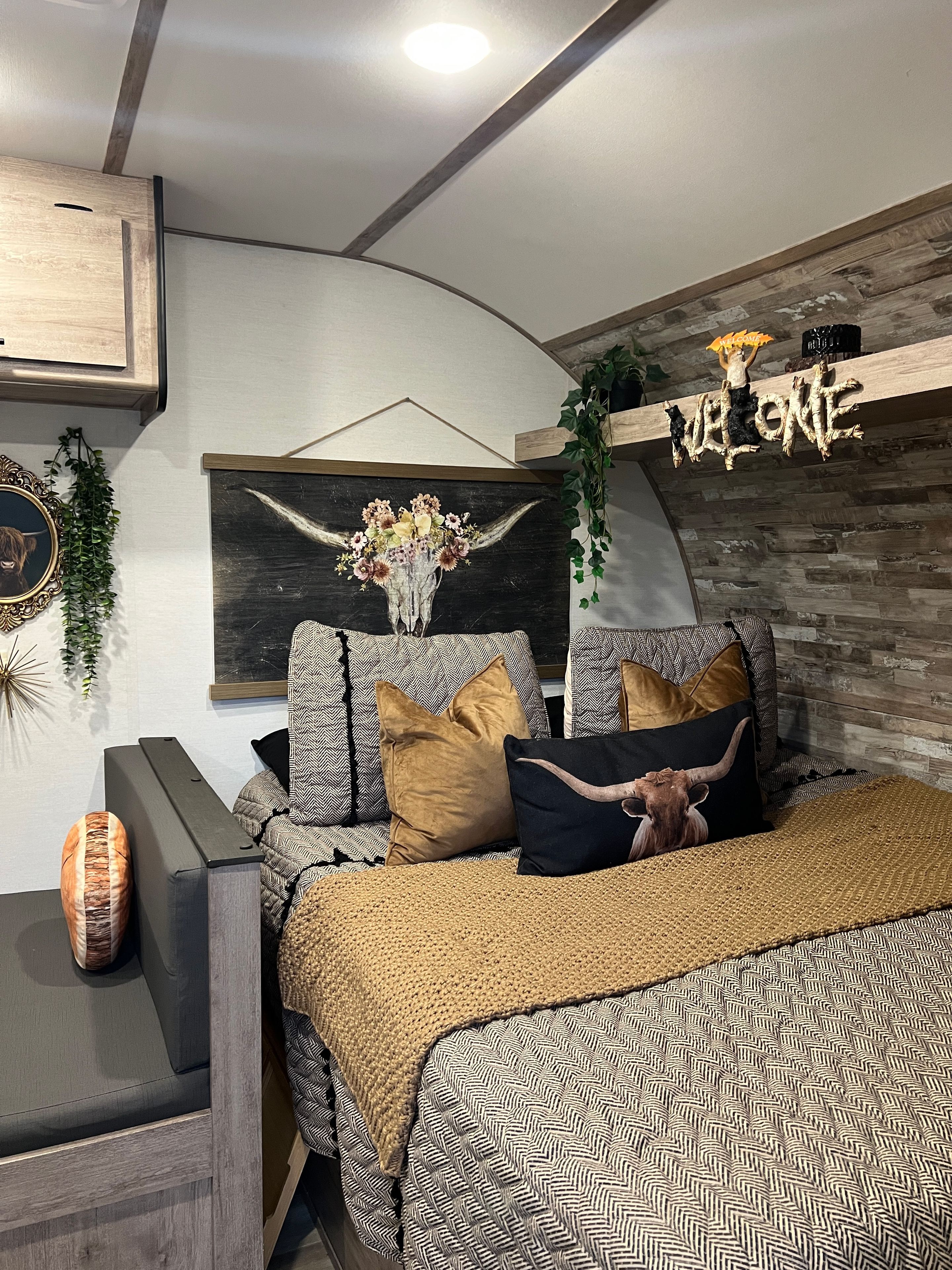 Un dormitorio con cama, almohadas, una obra de arte en la pared, un estante de madera con un letrero de bienvenida y una planta.
