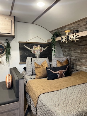A bedroom with a bed, pillows, a wall-mounted artwork, a wooden shelf with a welcome sign, and a plant.