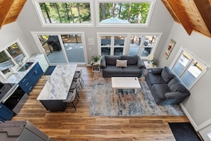A spacious living area with a kitchen, dining area, and a view of the outdoors through large windows. - 3-bed Maine Lakefront Home w/Private Dock & Fire Pit (Waterboro)