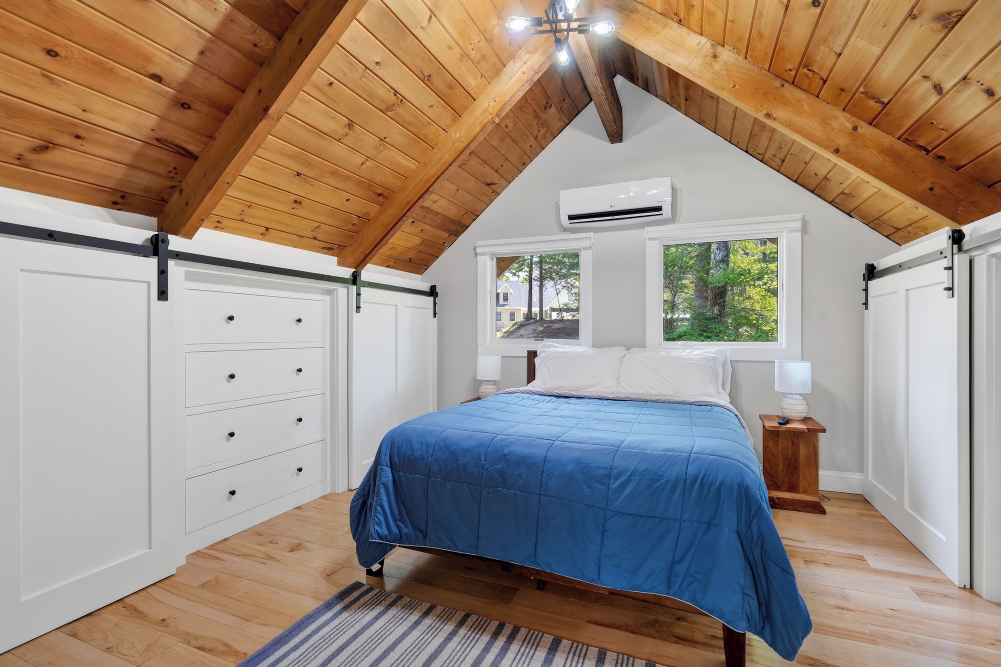 A bedroom with a sloped ceiling, a bed with a blue bedspread, a window with a view of trees, and an air conditioning unit.