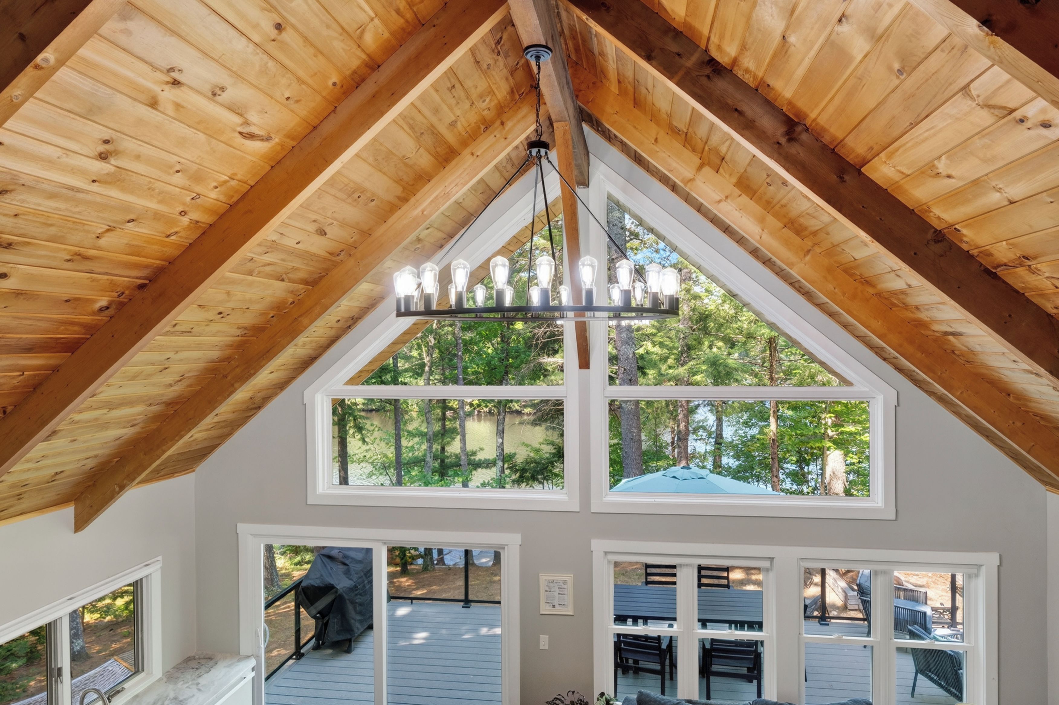 A room with a sloped wooden ceiling, a chandelier, and large windows offering a view of a deck and trees.