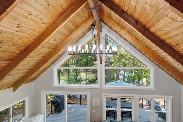 A room with a sloped wooden ceiling, a chandelier, and large windows offering a view of a deck and trees.