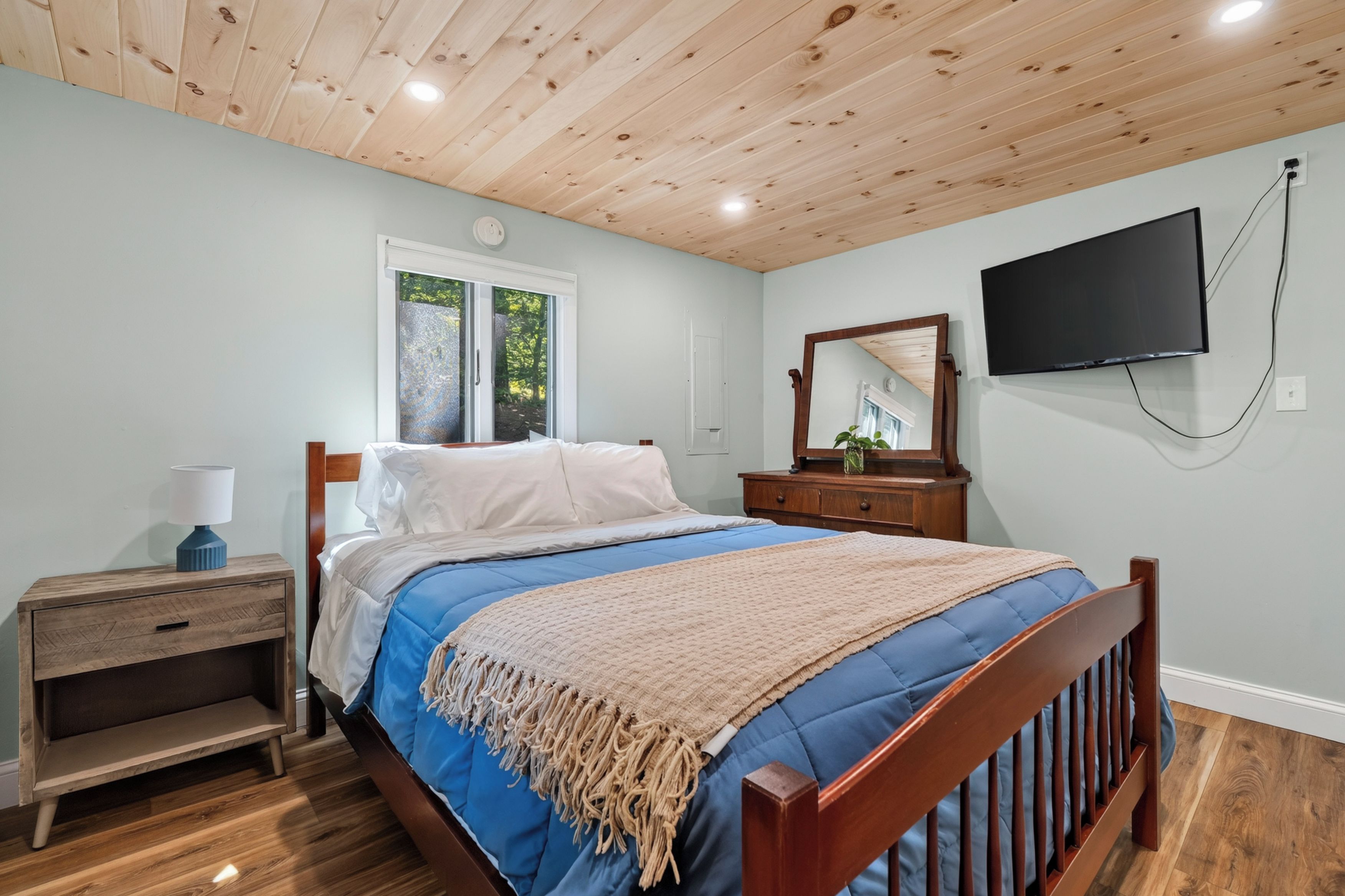 A bedroom with a wooden ceiling, a bed with blue bedding, a nightstand, a dresser, a mirror, and a flat-screen TV.