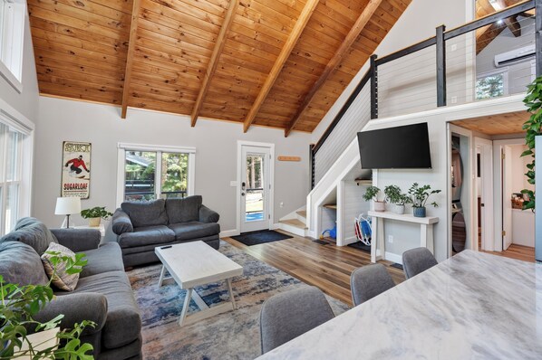 A spacious living room with a high wooden ceiling, a grey sofa set, a coffee table, and a TV mounted on the wall.