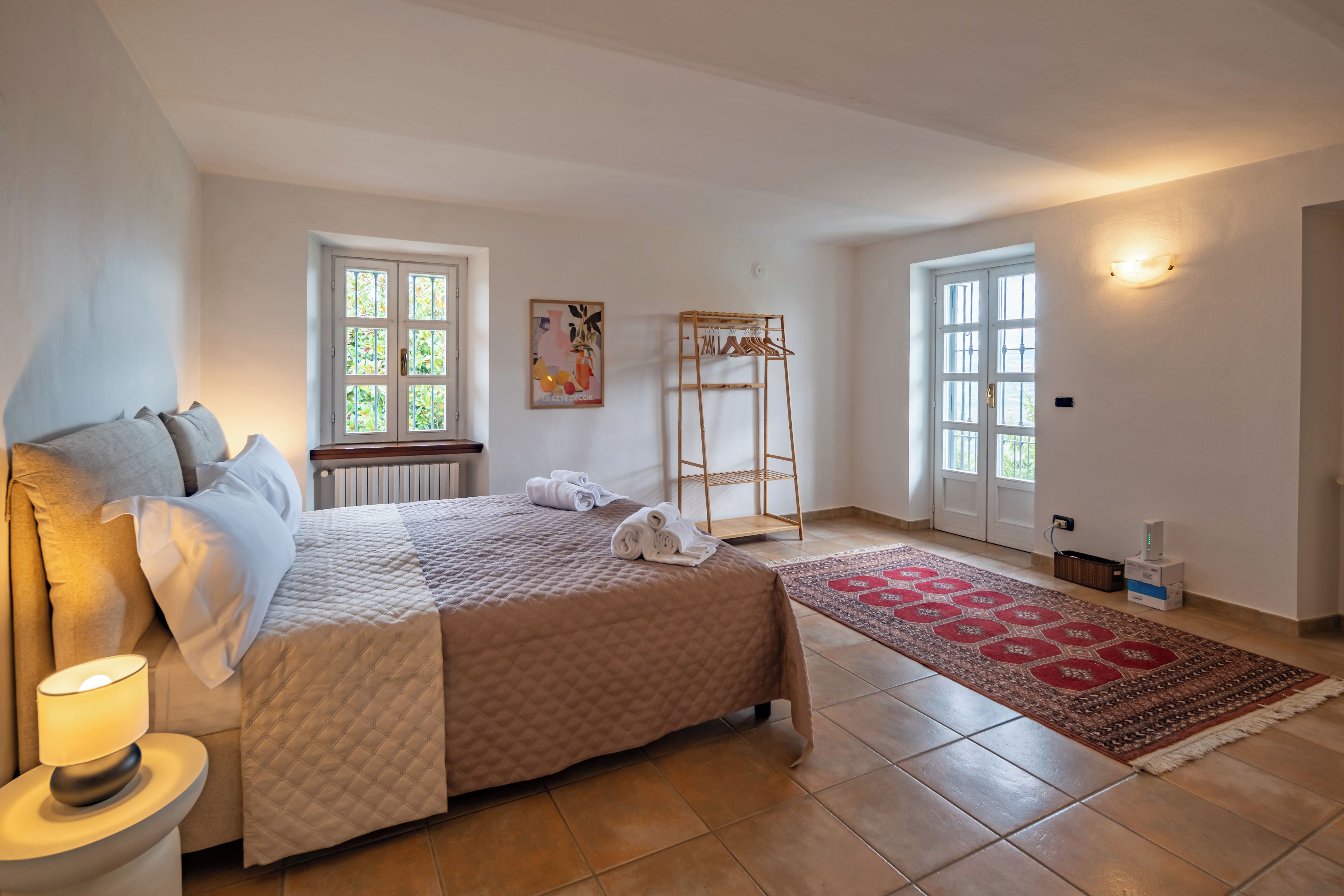 A bedroom with a bed, a bedside table, a window, a door, a wall-mounted light, and a rug.