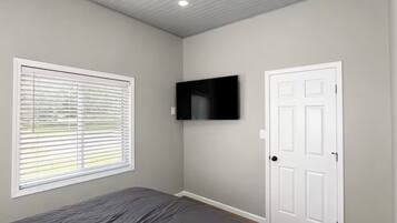 Ein Schlafzimmer mit einem Bett, einem an der Wand montierten Fernseher, einer Tür und einem Fenster mit Jalousien.