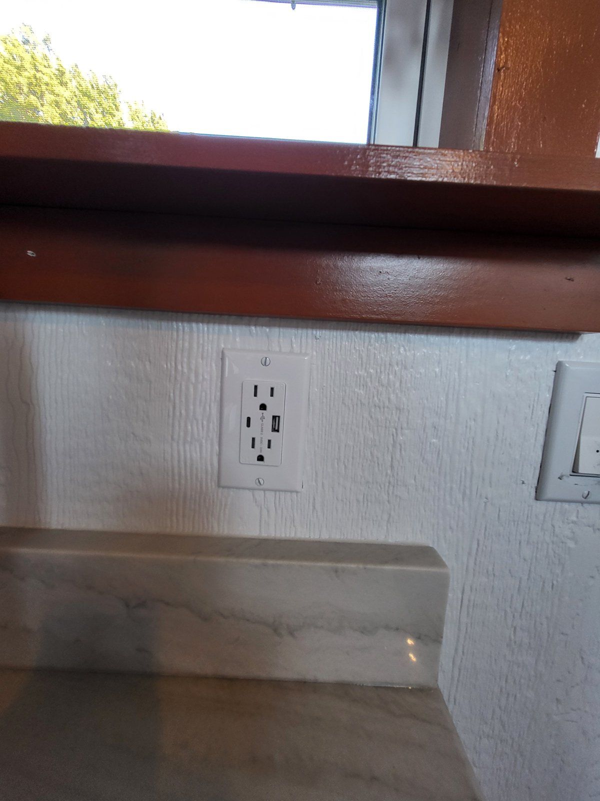 A white wall with a single electrical outlet and a window above it.