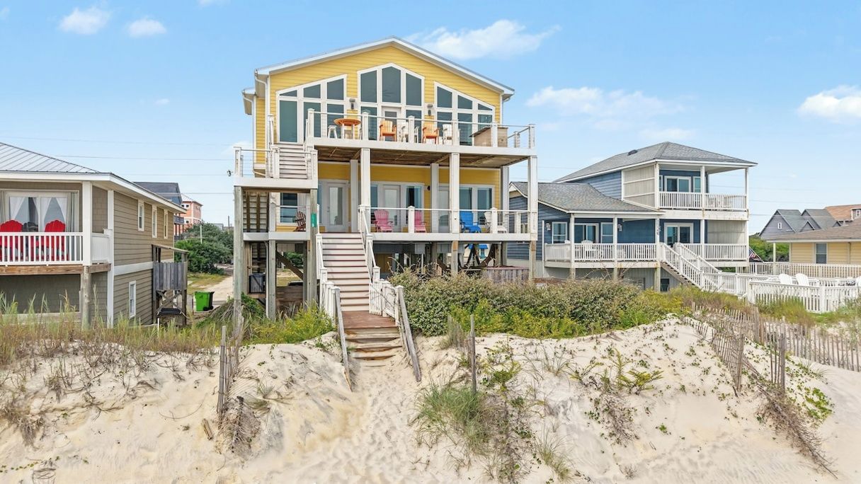 A multi-story beachfront house with a balcony, a staircase leading to the upper level, and a sandy beach in the foreground.