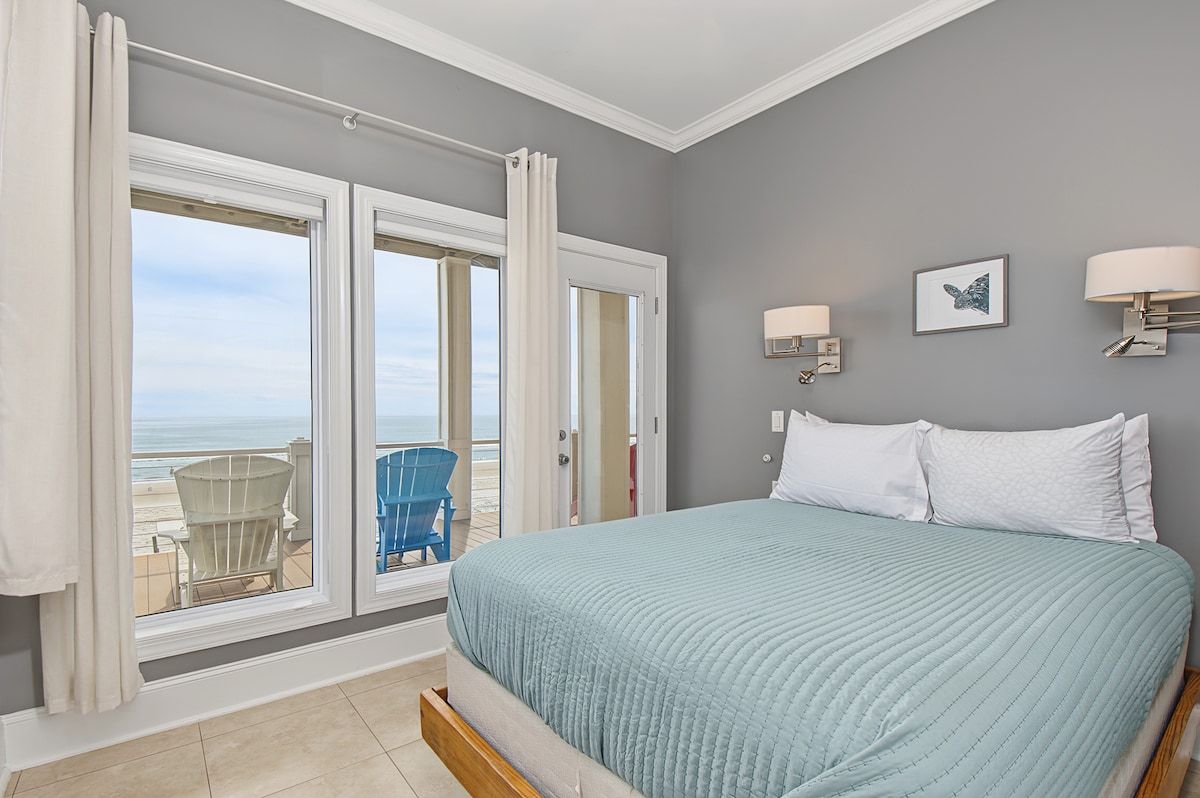 A bedroom with a bed, a view of the beach, and a chair outside the window.